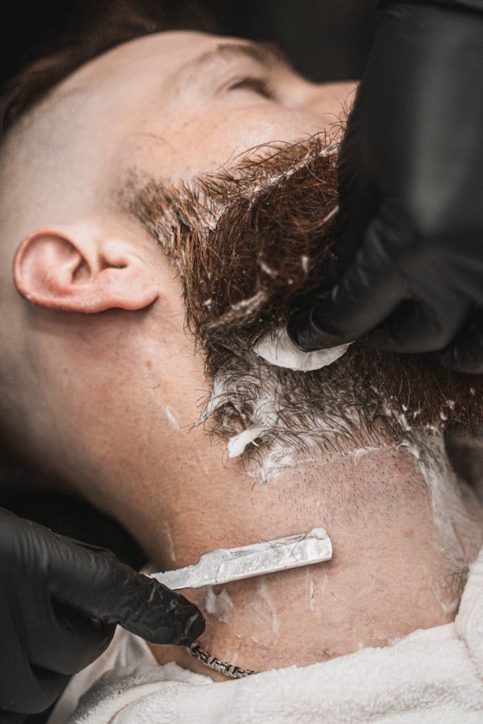 Barber expertly shaving a man's beard with a straight razor in a barbershop setting.