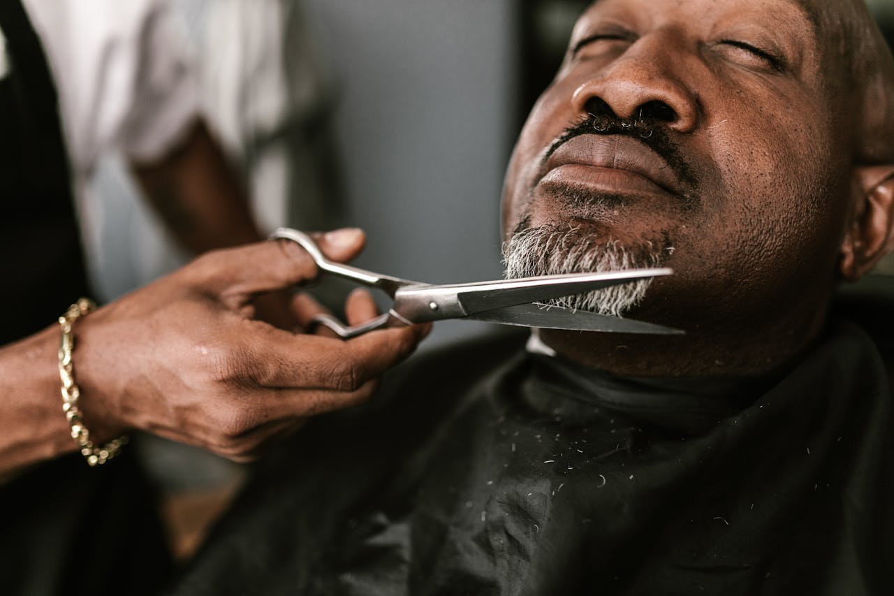 A professional barber skillfully trims a mature man's beard in a barbershop setting.