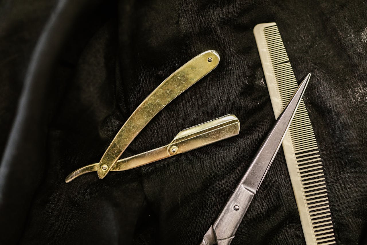 about-us Close-up of silver shears, comb, and straight razor on a black cloth background, highlighting barber tools.